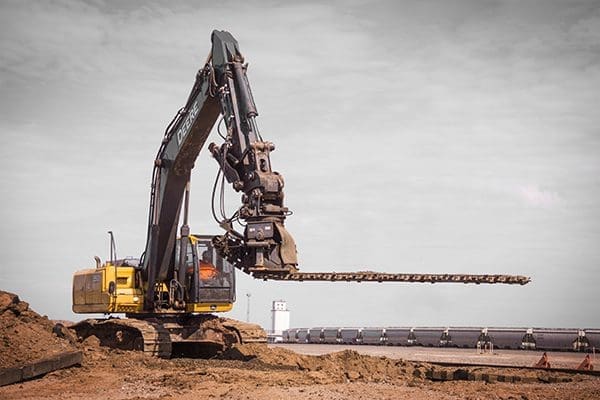 Ballast Blaster Undercutter – Large John Deere Excavator, Full Bar Extension DymaxRail Ballast Blaster undercutter mounted on large John Deere excavator with cutter bar at full horizontal extension, working next to excavated spoil pile at rail yard