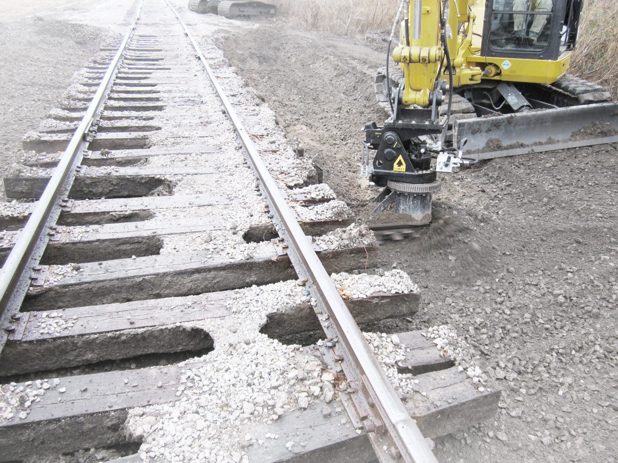 Ballast Blaster – Small Excavator Undercutting Between Ties, Exposed Crib DymaxRail Ballast Blaster undercutter on compact excavator cutting between railway ties, showing fouled ballast and mud removed from cribs with ties fully exposed