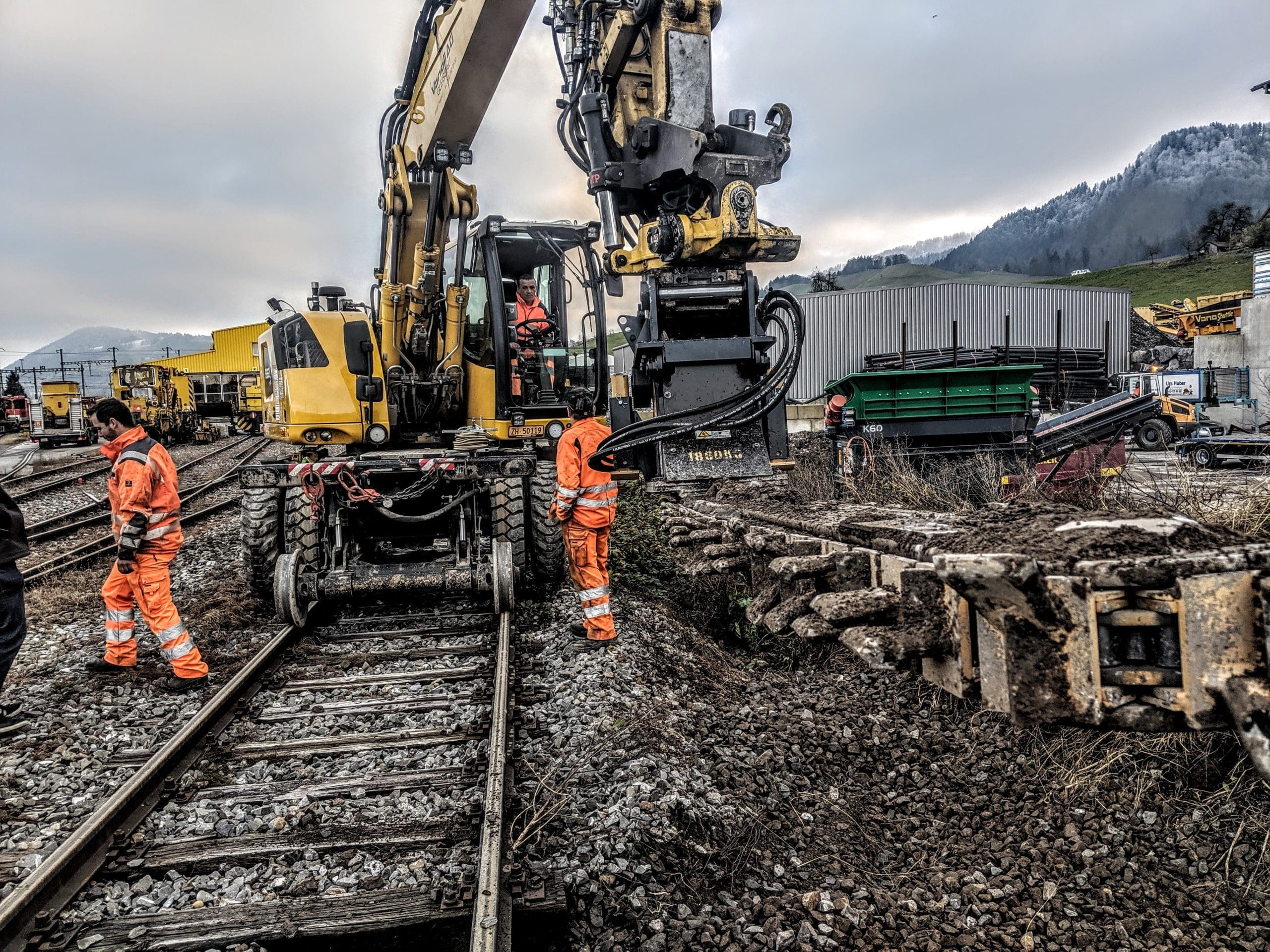 Ballast Blaster Tiltrotator – Active Fouled Ballast Removal with Crew DymaxRail Ballast Blaster tiltrotator undercutter removing heavily fouled ballast and mud from beneath railway track, operator in cab with ground crew in hi-vis working alongside