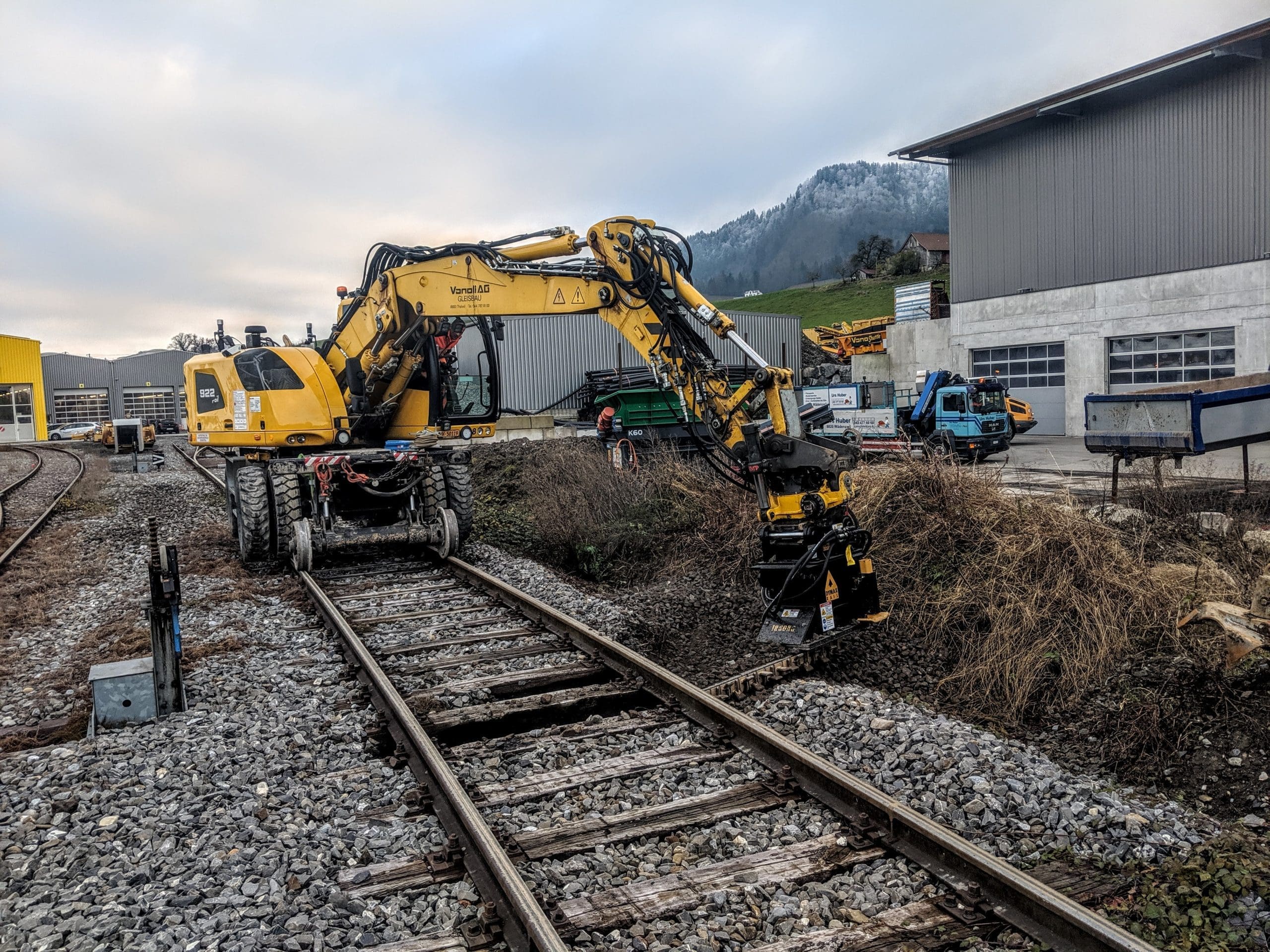 Ballast Blaster Tiltrotator – Wheel Excavator on Hi-Rail, Working from Track Side DymaxRail Ballast Blaster tiltrotator undercutter mounted on hi-railed wheel excavator, positioned to cut from the side of the railway track during ballast maintenance