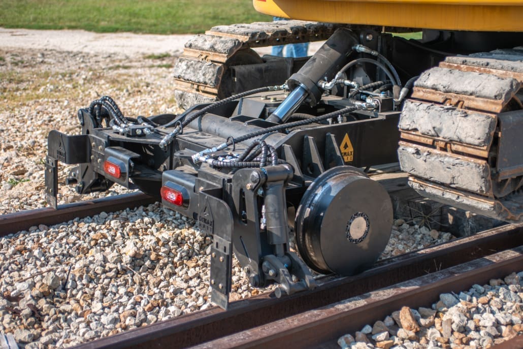 Dymax Rail Rider 8T hi-rail front railgear detail on rail Detail view of the Dymax Rail Rider 8T hi-rail front railgear seated on rail beneath a Cat track excavator, showing rail wheels, hydraulic clamps, and LED lighting modules