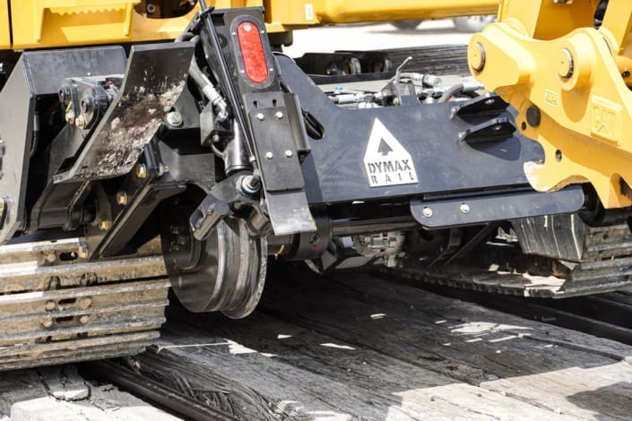 Dymax Rail Rider X2-15T with 16-inch rail wheels Close-up of the Dymax Rail Rider X2-15T hi-rail undercarriage on wood ties, showing 16-inch steel rail wheel and replaceable brake shoe on a Cat track excavator