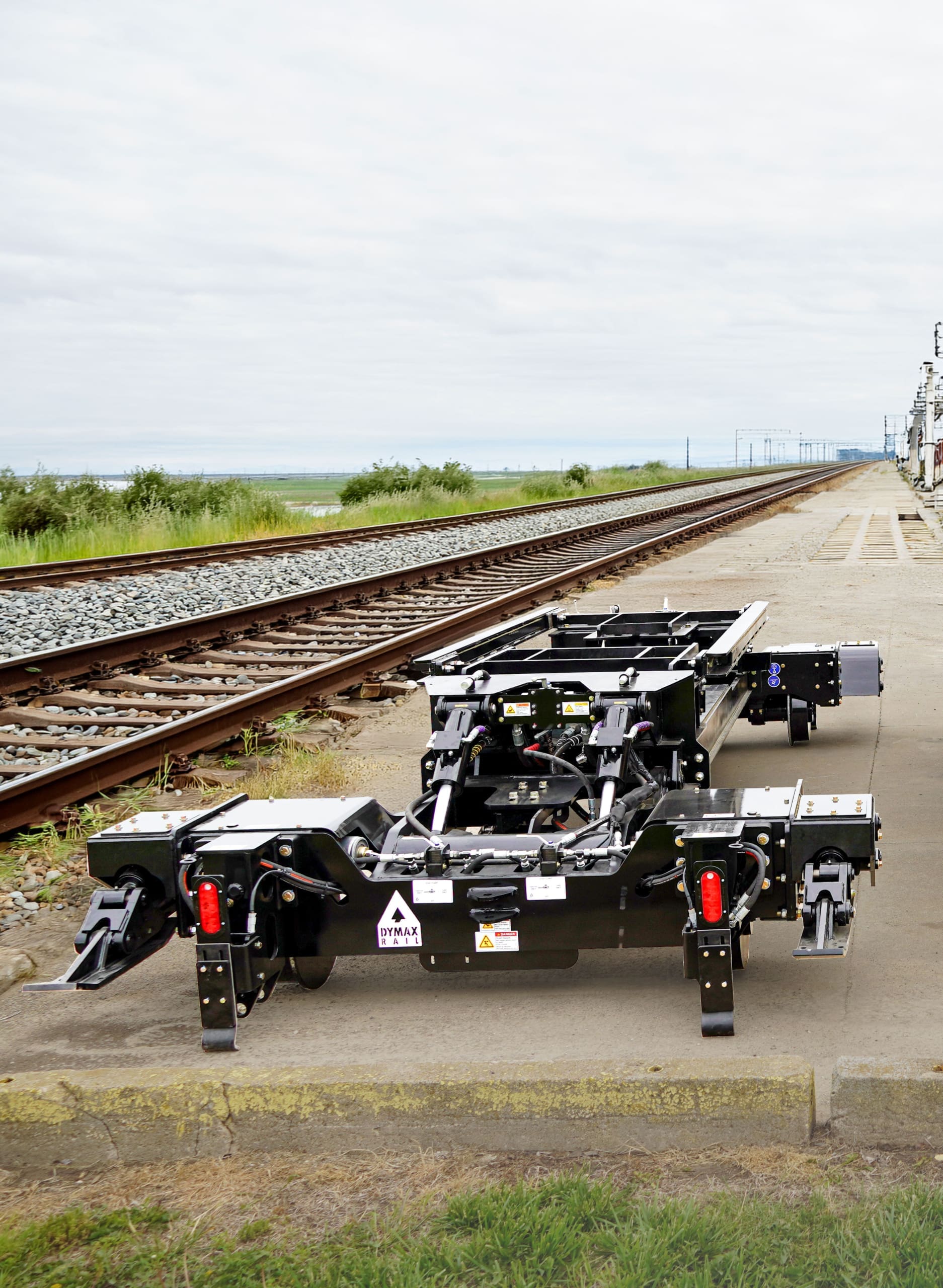 Dymax Rail Rider X2 detachable hi-rail attachment shown standalone on a concrete pad next to an active rail line, ready for installation on a track excavator