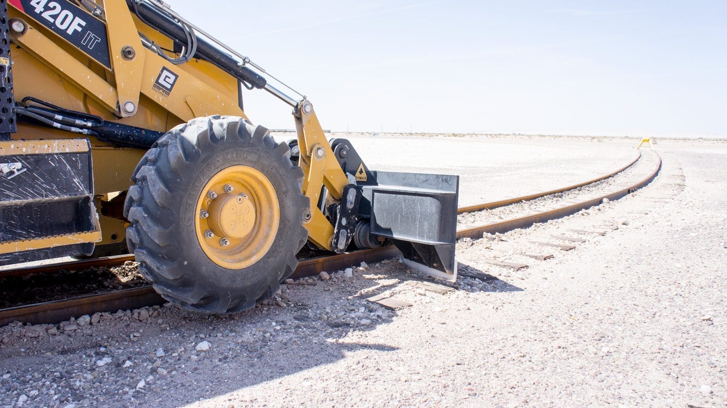 DymaxRail Rail Ballast Plow attached to Cat 420F backhoe loader, guided on rail in yard setting