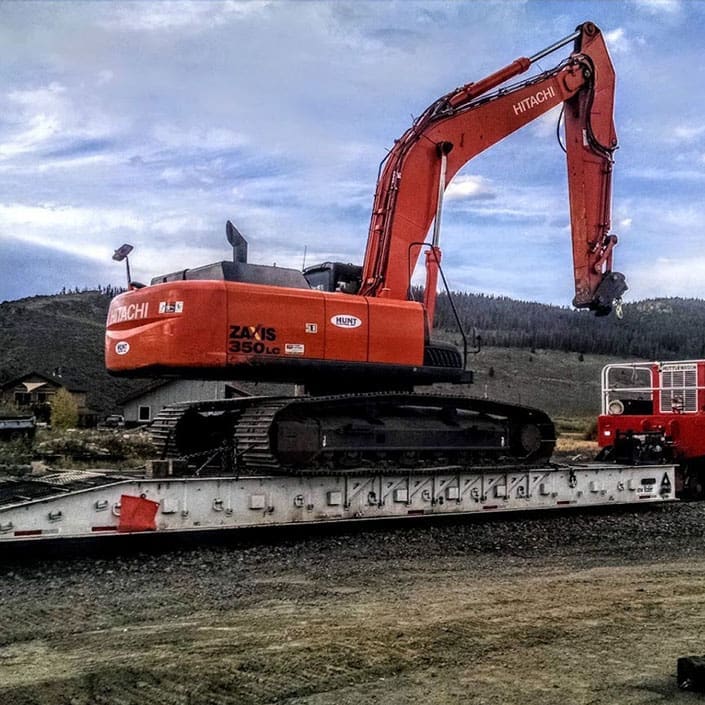 Machinery Cart - Towable Large excavator being transported on rail by a machinery rail cart and Shuttlewagon railcar mover, with mountains and blue sky in background.