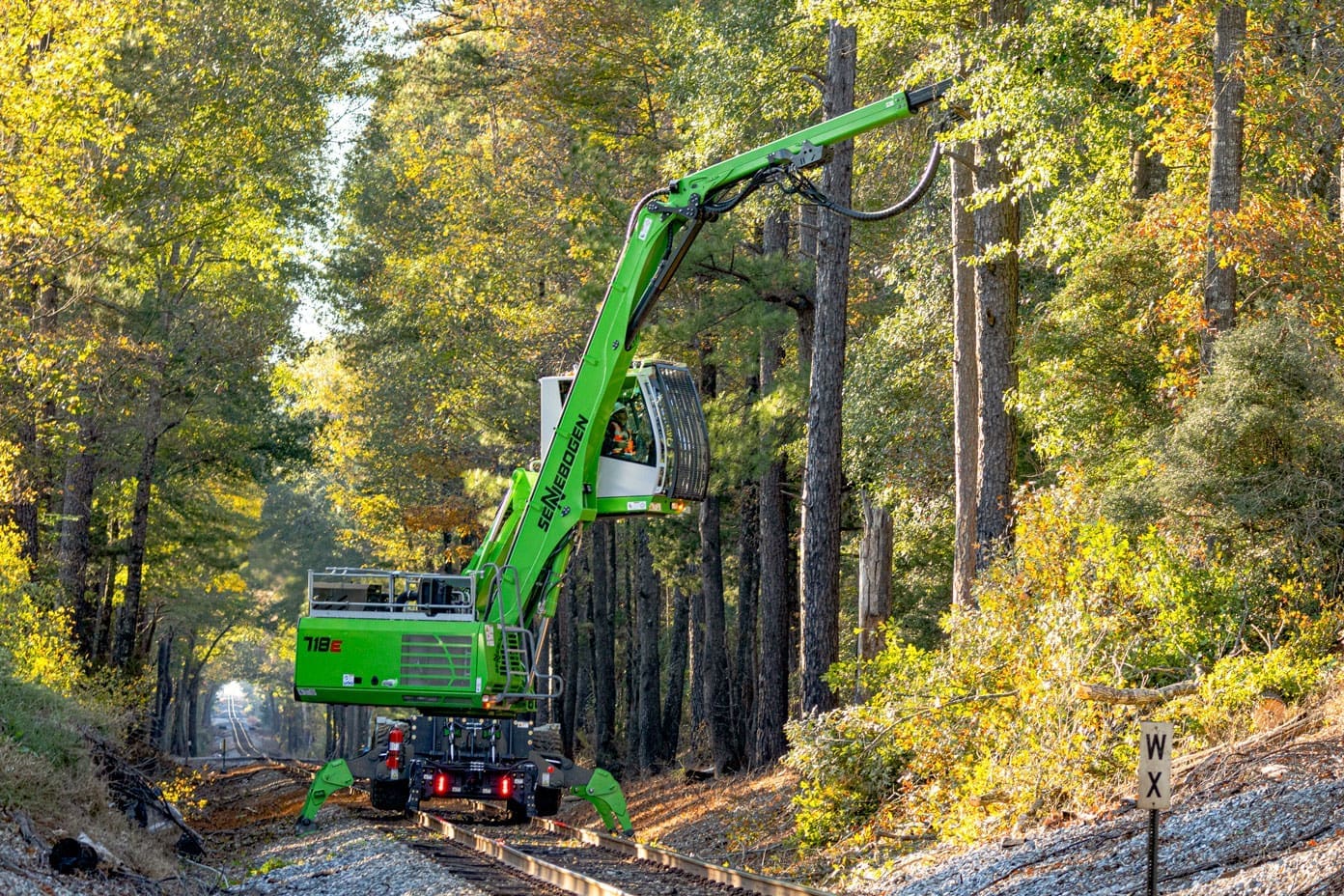 dymax-rail-rider-x2-hi-rail-track-excavator-rail-conversion Track excavator converted with Dymax Rail Rider X2 detachable hi-rail system operating on railroad track in forested area